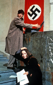 Szenenbild aus dem Drama "Die weiße Rose" (1982): Ein junger Mann hält steht auf einer Treppe und hält einen Koffer auf, der auf einen Vorsprung steht. Er schaut sich um. Neben ihm kniet auf der Treppe eine junge Frau. Sie hält Papierblätter in der Hand. Beide wirken angespannt. An der Wand hängt eine Hakenkreuz-Flagge. (© picture alliance)