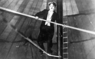 Schwarz-weißes Szenenbild aus dem Stummfilm Der Zirkus (USA 1927): Der Schauspieler Charlie Chaplin balanciert in einem Zirkuszelt auf einem Seil und hält ein lange Stange in den Händen. (© picture-alliance / akg-images | akg-images)