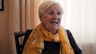 Ihr Jahrhundert – Frauen erzählen Geschichte, Szenenbild: Eine alte Frau mit fast weißen Haaren sitzt auf einem Stuhl und spricht. Sie trägt ein gelbes Tuch um den Hals. (© Mindjazz Pictures)
