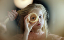 Romys Salon, Szenenbild: Naheinstellung: Ein blondes Mädchen mit einem Frisierumhang schaut direkt in die Kamera und hält sich einen Lockenwickler vor ein Auge. (© Elmer van der Marel/Farbfilm Verleih)