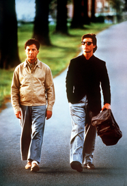 Rain Man, Szenenbild: Zwei Männer, einer ist älter, laufen nebeneinander eine Straße lang. Der jüngere Mann trägt eine Sonnenbrille und eine große Tasche. (© United Artists/picture alliance/dpa)