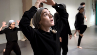 1001 Nights Apart, Szenenbild: Eine junge, schwarz gekleidete Tänzerin mit Kopftuch hält ihre Arme über ihren Kopf und schaut nach oben. Im Hintergrund sind weitere Frauen und Männer bei einer Tanzprobe zu sehen. (© Filmpunkt)