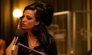 Szenenbild aus dem Film &quot;Back to Black&quot;: Die Sängerin Amy Winehouse, dargestellt von Marisa Abela, steht in einem Tonstudio vor einem Mikrofon und singt. Sie hält sich einen Kophörer an ihr rechtes Ohr. (© Dean Rogers / Focus Features)
