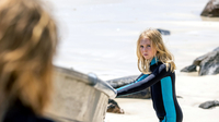 Blueback - Eine tiefe Freundschaft, Szenenbild: Ein Kind steht im Taucheranzug am Strand und hält sich am Rand eines Bootes fest. Es schaut mit ernstem Blick in Richtung einer Person, von der im Vordergrund nur der Hinterkopf unscharf zu erkennen ist (© Arenamedia Pty Ltd, Foto: Nic Duncan)