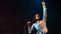 Bob Marley: One Love, Szenenbild: Ein Schwarzer Musiker mit Rastalocken steht auf einer Bühne an einem Mikrofonständer. Er hebt einen Arm gerade nach oben. (© Paramount Pictures)