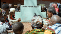Szenenbild aus dem Dokumentarfilm "Das leere Grab": Nahaufnahme eines Grabstein mit der Inschrift "Nduna Songea Mbano". Um den Stein knien Frauen und Männer. Das Bild wurde in Tansania aufgenommen.(© Salzgeber)