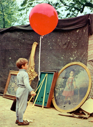 Der rote Ballon, Szenenbild: Ein kleiner, grau gekleideter Junge steht mit einem großen, roten Luftballon vor einem Gemälde. Es zeigt ein Mädchen mit einem Reifen. (© picture alliance/Everett Collection)