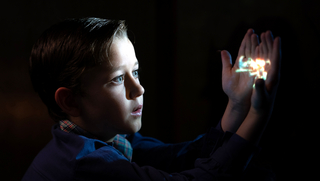 Die Fabelmans, Szenenbild: Ein kleiner Junge, seitlich aufgenommen, sitzt im Dunkeln. Sein Gesicht ist von rechts angestrahlt. Er hält seine Hände vor sich, auf die ein Filmbild projiziert ist. (© Storyteller Distribution Co., LLC. All Rights Reserved)