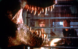 The Lost World: Jurassic Park, Szenenbild: Großaufnahme eines Tyranosaurus Rex mit geöffneten Maul, im Hintergrund sind zwei Menschen zu sehen. (© picture-alliance / dpa | dpa-Film)