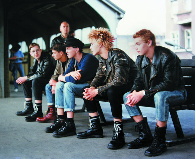 Flüstern & Schreien, Szenenbild: Fünf junge Männer in Jeans und Springerstiefeln sitzen nebeneinander auf einer Bank auf einem Bahnsteig. (© DEFA Stiftung, Tina Bara)