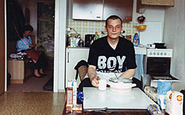 Stau – Jetzt geht's los, Szenebild: Ein junger Mann sitzt an einem Tisch in der Küche. Auf seinem T-Shirt steht &quot;Boy&quot;. Im Hintergrund sitzt die Mutter in einem anderen Zimmer. (© ÖFilm Dörr + Schlösser GmbH)