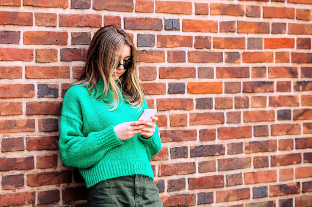 Ein Mädchen im Teenageralter lehnt an einer Mauer und schaut auf ihr Handy (© Westend61 / Vira Simon / picture alliance)