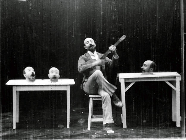 Szenenbild aus dem Film Un homme de têtes von Georges Méliès (1898): Ein Mann sitzt mit einer Gitarre auf einem Stuhl. Auf Tischen neben ihm liegen drei Köpfe von ihm, die singen. (© StudioCanal/Arthaus)