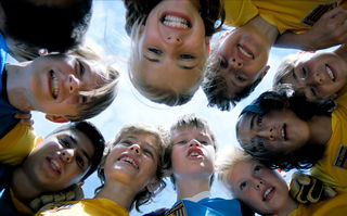 Zu weit weg, Szennebild: Von unten aufgenommen: Mädchen und Jungen im Fußballtrikot, die sich im Kreis umarmen und lachen (© Weydemann Bros. GmbH / Monika Plura)