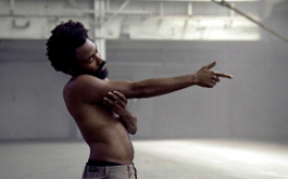 This is America, Szenenbild: Nahaufnahme des afroamerikanischen Musikers Childish Gambino. Er steht mit nacktem Oberkörper in einer Halle und zielt mit dem Finger nach rechts. (© Hiro Murai)