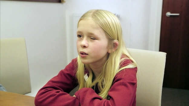 Nora in New York, Szenenbild (© WDR): Nora, 13 Jahre alt, sitzt auf einem Stuhl und blickt an der Kamera; ihr linkes Auge, auf dem sie nicht sehen kann, ist geschlossen.
