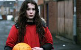 Der Ball, Szenenbild: Nahaufname eines dunkelhaarigen Mädchens in einem roten Pullover. Es steht in einer Gasse und hält einen Ball in den Händen. (© Merkaat Films)