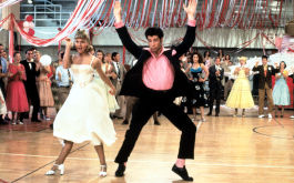 Grease, Szenenbild: Eine blonde Frau im weißen Kleid und ein junger Mann im schwarzen Anzug und rosa Hemd tanzen ausgelassen in einer Turnhalle (© picture alliance / ZUMAPRESS.com | Paramount Pictures)