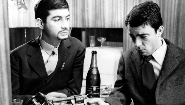 Schrei, wenn du kannst, Szenenbild: Zwei junge Männer stehen nebeneinander. Sie schauen ernst. Zwischen ihnen steht eine Flasche Champagner. (© ddp images)