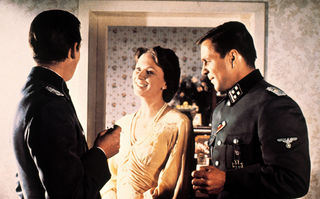 Aus einem deutschen Leben, Szenenbild: Zwei Männer in SS-Uniformen und eine Frau im hellen Kleid stehen zusammen in einem Gespräch. Die Frau lächelt. (© picture alliance/United Archives)
