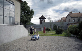 The Zone of Interest, Szenenbild: Zwei Frauen und zwei Kinder stehen neben einem Wohnhaus in einem Garten, der von einer hohen Mauer begrenzt ist. Im Hintergrund sind Gebäude und der Wachturm eines Konzentrationslagers zu sehen. (© Leonine Studios)