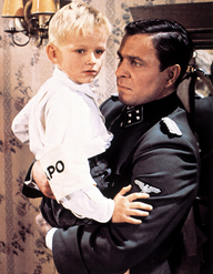 Aus einem deutschen Leben, Szenenbild: Ein Mann in SS-Uniform trägt einen kleinen, blonden Jungen im Arm und schaut ihn an. Der Junge trägt eine Armbinde, auf der "apo" ("Gestapo") zu lesen ist.(© picture alliance/United Archives | United Archives / kpa Publicity)