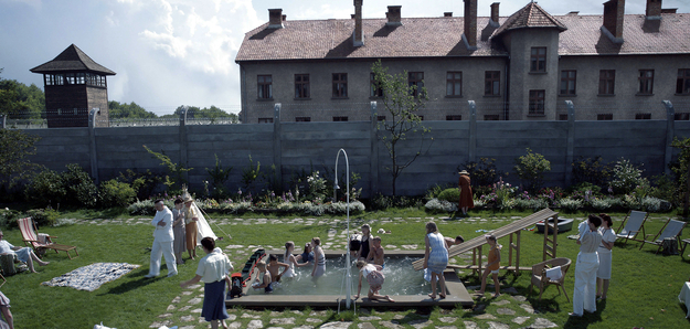 The Zone of Interest, Szenenbild: Menschen und Kinder halten sich in einem sommerlichen Garten mit Planschbecken auf. Der Garten ist von einer hohen Mauer begrenzt. Dahinter ist eine Haus und der Wachturm eines Konzentrationslagers zu erkennen. (© Leonine Studios)