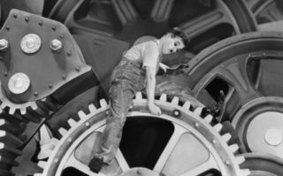 Schwarz-weißes Szenenbild aus dem Film Moderne Zeiten (USA 1936): Der Schauspieler Charlie Chaplin sitzt auf einem riesigen Zahnrad und hat ein Werkzeug in der Hand. Er trägt Arbeitskleidung. (© picture alliance)