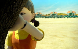 Squid Game, Szenenbild: Im Vordergrund steht am linken Bildrand eine übermenschlich große Puppe, die an einen Baum gelehnt ist. Im Hintergrund stehen weit entfernt grün gekleidete Menschen. (© Netflix)