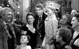 Ist das Leben nicht schön? Schwarz-weißes Szenenbild: Ein Gruppe von Menschen steht neben einem geschmückten Weihnachtsbaum und hört einem am linken Bildrand stehenden Mann zu. (© Masheter Movie Archive / Alamy Stock Photo)
