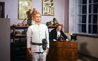 Woyzeck, Szenenbild: Ein kahlköpfiger Mann in einem einfachen, weißen Anzug steht in einem Büro stramm. Hinter einem Schreibtisch beugt sich lächelnd ein Mann vor. (© Werner Herzog Film)