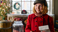 Ein Weihnachtsfest für Teddy, Szenenbild: Ein lächelndes rothaariges Mädchen mit Mütze und in einem roten Mantel steht mit einer Papiertüte in der Hand in einem weihnachtlich dekorierten Laden (© Capelight Pictures)