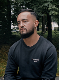 Halbnahe Einstellung eines ins Leere blickenden jungen Mannes in einem schwarzem Sweatshirt vor Bäumen (© ZDF/Julian Vogel)
