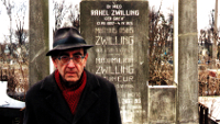 Herr Zwilling und Frau Zuckermann, Szenenbild: Ein älterer Herr steht auf einem jüdischen Friedhof vor dem Grab von Angehörigen. Er schaut direkt in die Kamera. (© Editiona Salzgeber)