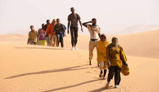 Szenenbild aus dem film &quot;Ich Capitano&quot;: Eine Reihe von Schwarzen Frauen und Männern laufen im Gänsemarsch durch eine Wüstenlandschaft. (© Greta De Lazzaris / X Verleih AG)