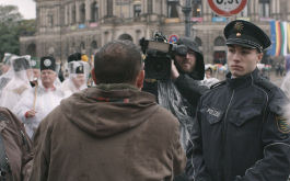 Aggregat, Szenenbild: Eine Pegida-Demonstration in Dresden: Von hinten ist ein Mann zu sehen, der vor einem Medienvertreter mit Kamera steht, zwischen ihnen ein junger Polizist. (© Zorro Film)