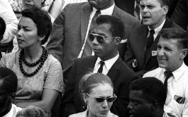 I Am Not Your Negro, Szenenbild: Halbnahe Einstellung eines Publikums. In der Mitte sitzt der afroamerikanische Schriftsteller James Baldwin mit Sonnenbrille. Das Bild stammt aus de 1960ern. (© Edition Salzgeber)
