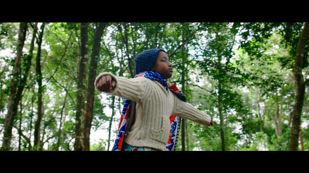 Supa Modo, Szenenbild: Das Mädchen Jo steht mit ausgebreiteten Armen und geschlossenen Augen in einem Wald. Sie trägt eine blaue Wollmütze, eine weiße Strickjacke und einen bunten Umhang . (© One Fine Day Films)