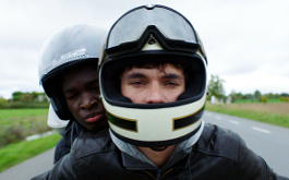 Roads, Szenenbild: Nahaufnahme, frontal: Der 18-jährige Brite Gyllen und der fast gleichaltrige Kongolese William fahren auf einem Mottorrad. Sie Tragen Helme (© Studiocanal GmbH) 