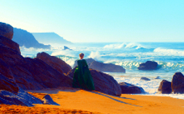 Porträt einer jungen Frau in Flammen, Szenenbild: Totale, Rückenansicht: Eine junge blonde Frau im langen Kleid steht am Strand und blickt aufs Meer. Am linken Bildrand befindet sich eine Klippe. (© Alamode Film)