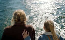 Romys Salon, Szenenbild: Rückenansicht eines blonden Mädchens und einer älteren Frau, die gemeinsam auf das Meer schauen. Das Kind hat ihre Hand auf den Rücken der Frau gelegt. (© Melle van Essen/Farbfilm Verleih)