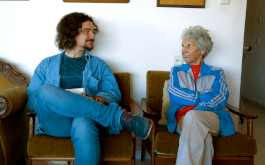 Endlich Tacheles, Szenenbild: Ein junger Mann und eine alte Damen setzen nebeneinander auf Stühlen und schauen sich an. (© Schramm Matthes Film)