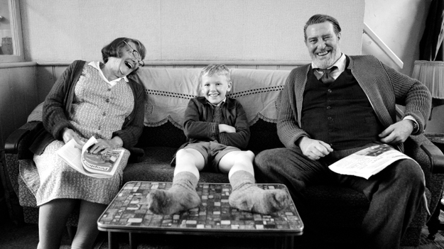 Belfast, Szenenbild: Ein Junge sitzt auf einem Sofa, Füße auf den Tisch. Neben ihm sind seine Großeltern. Alle lachen. (© Rob Youngson / Focus Features)