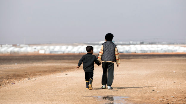 Totale: Zwei Flüchtlingskinder in der Rückenansicht laufen Hand in Hand durch eine karge, sandige Landschaft. Im Hintergrund sind schemenhaft weiße Zelte zu erkennen. Das Foto wurde am 29.01.2018 in Jordanien im Flüchtlingslager Al-Asrak aufgenommen. (© picture alliance / Jörg Carstensen/dpa)