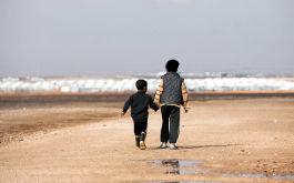 Totale: Zwei Flüchtlingskinder in der Rückenansicht laufen Hand in Hand durch eine karge, sandige Landschaft. Im Hintergrund sind schemenhaft weiße Zelte zu erkennen. Das Foto wurde am 29.01.2018 in Jordanien im Flüchtlingslager Al-Asrak aufgenommen. (© picture alliance / Jörg Carstensen/dpa)