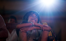 Das Licht, aus dem die Träume sind, Szenenbild: Eine Junge sitzt lächelnd und mit angezogenen Beinen in einem dunklen Kinosaal. Im Hintergrund sieht man das Projektorlicht. (© Neue Visionen)