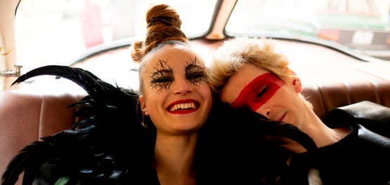 In einem Land, das es nicht mehr gibt, Szenenbild: Eine junge Frau mit schwazer Federboa und exzentrischem Make-up und eine Mann, mit einem aufgemalten, roten Streifen über den Augen sitzen lachend auf dem Hintersitz eines Autos. (© Ziegler Film/TOBIS/ Peter Hartwig)