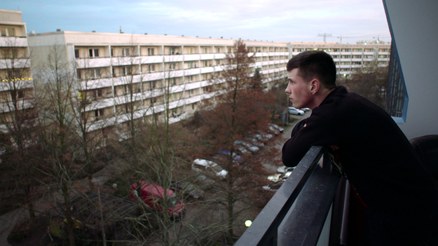 Kalle Kosmonaut, Szenenbild: Ein junger Mann steht am rechten Bildrand auf einem Balkon. Er schaut auf eine Straße mit Plattenbauten. (© Günther Kurth / mindjazz pictures)