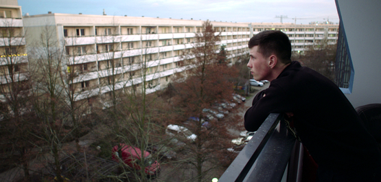 Kalle Kosmonaut, Szenenbild: Ein junger Mann steht am rechten Bildrand auf einem Balkon. Er schaut auf eine Straße mit Plattenbauten (© Günther Kurth / mindjazz pictures)