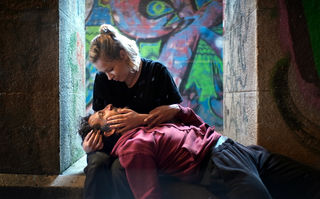 Orphea in Love, Szenenbild: Vor einer mit Graffitis besprühten Wand in einer Unterführung, hält eine Frau einen liegenden Mann in ihren Armen und schaut ihn an. (© missingFILMs)